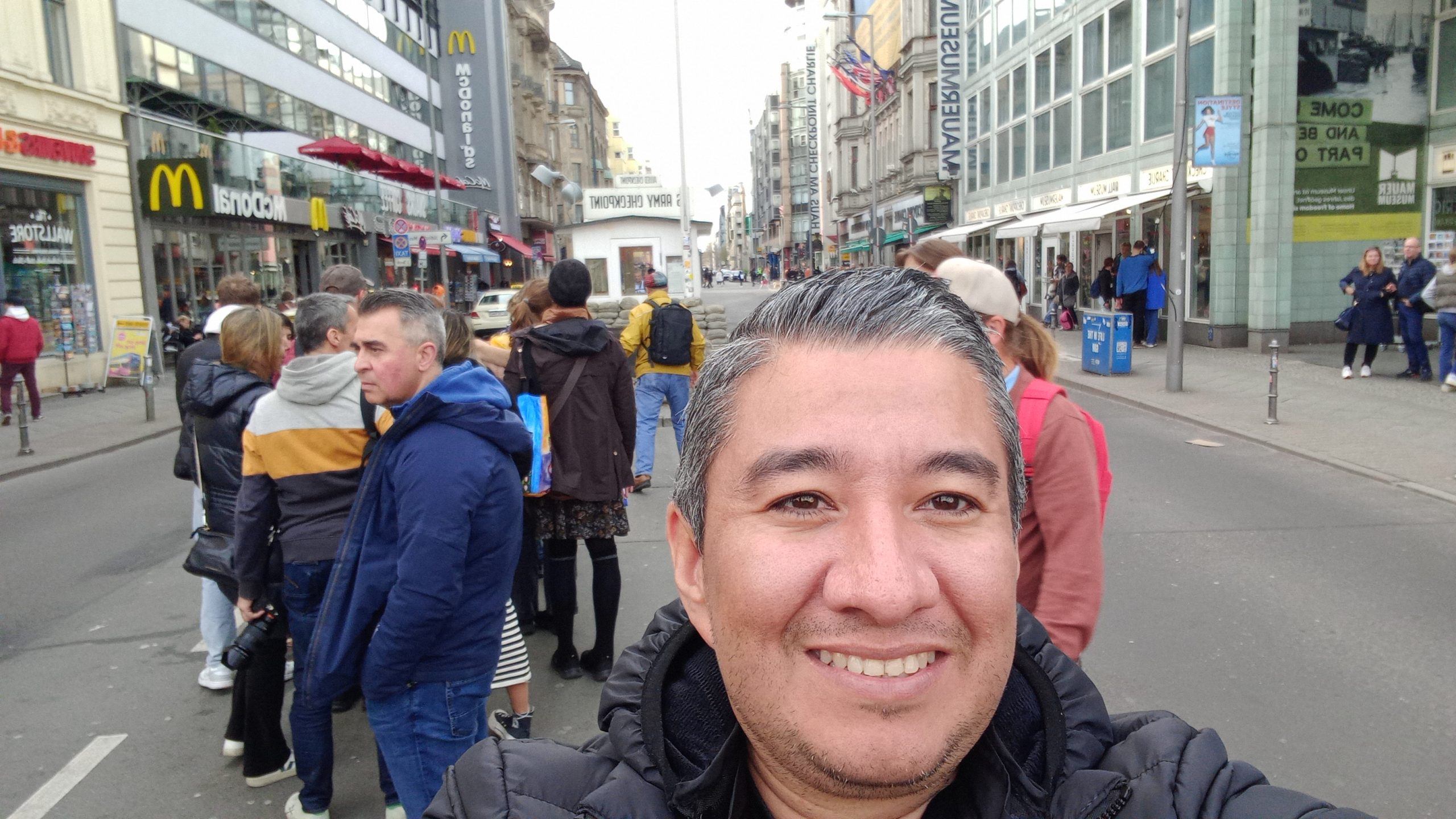 Fotografía frente a Checkpoint Charlie en Berlín, con McDonald’s y KFC al fondo, mostrando el contraste entre la Guerra Fría y la ciudad actual.