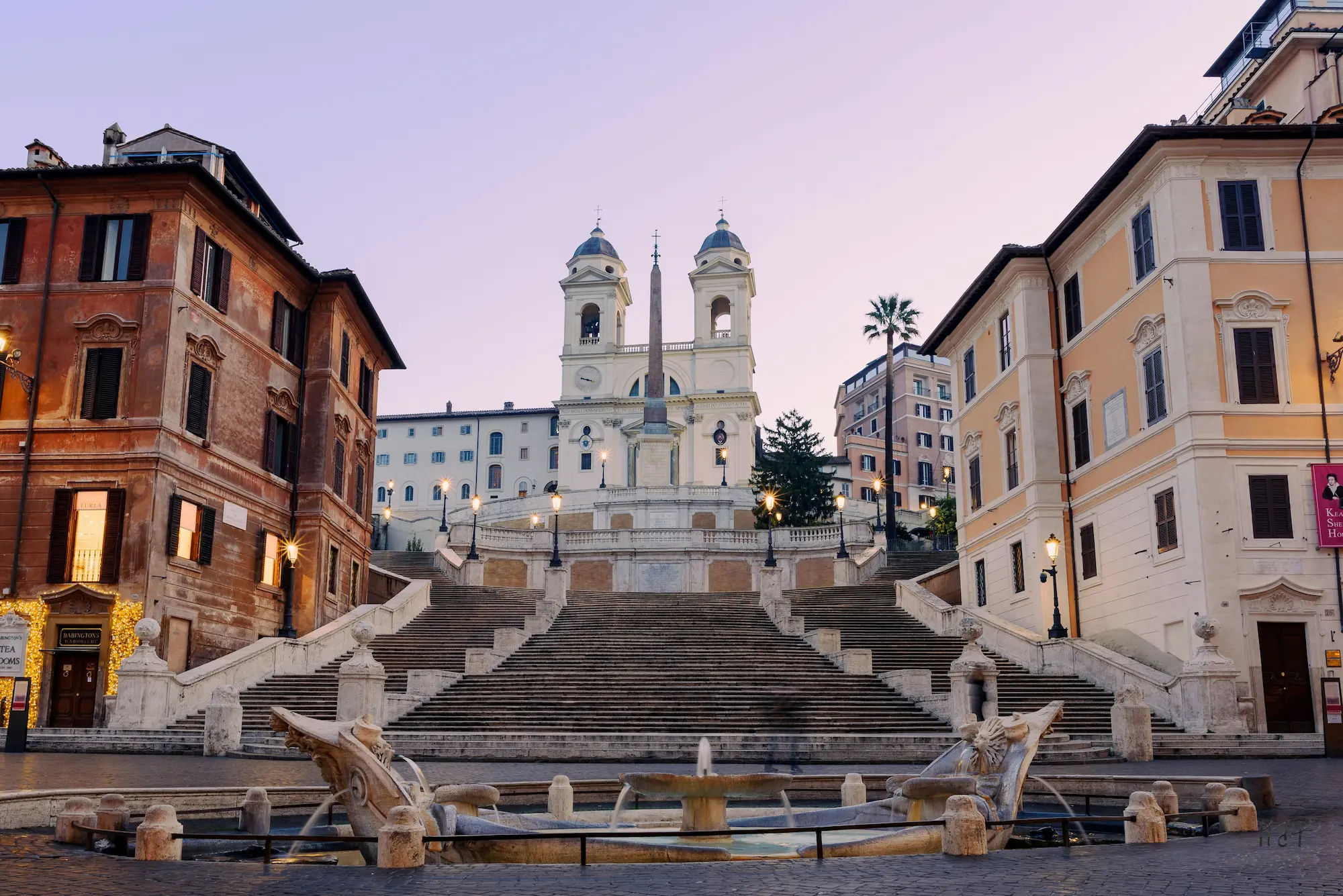 Vista frontal de la Escalinata de la Plaza de España en Roma al amanecer, con la Fuente de la Barcaza en primer plano y la iglesia de la Trinità dei Monti al fondo bajo un cielo rosado.
