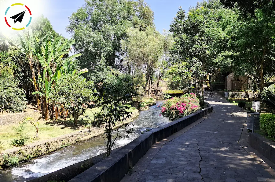 Sendero de piedra del Parque Lineal La Camelina en Uruapan, Michoacán, corriendo paralelo al río Cupatitzio con puentes rústicos y vegetación exuberante.