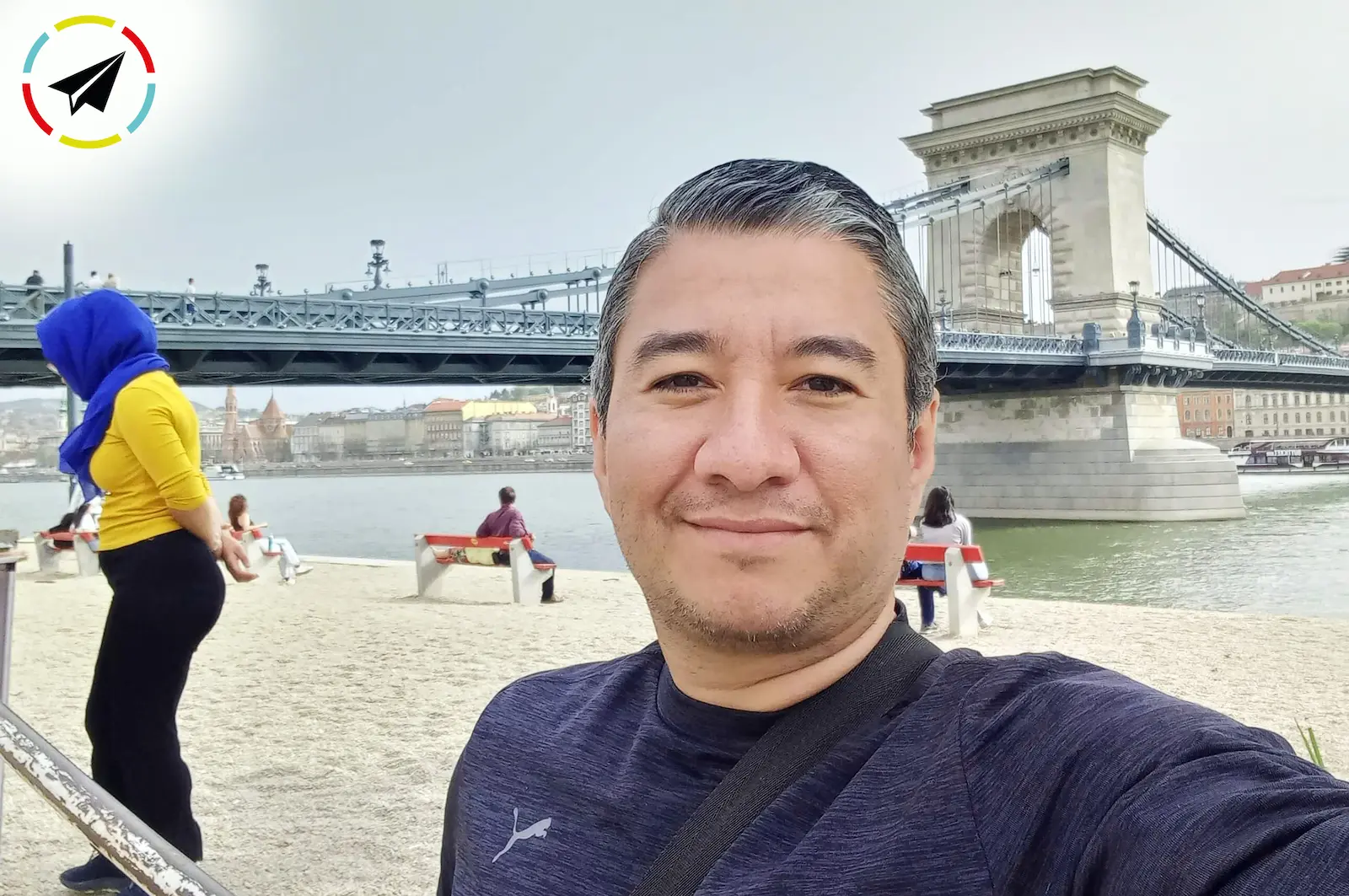 Parado en la orilla del río Danubio en Budapest, Hungría, con un puente metálico al fondo.