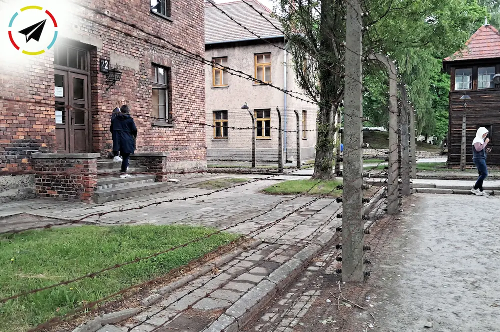 Cerca de alambre de púas electrificado rodeando el perímetro de Auschwitz-Birkenau, con una torre de vigilancia de ladrillo de los SS al fondo controlando todo el movimiento.