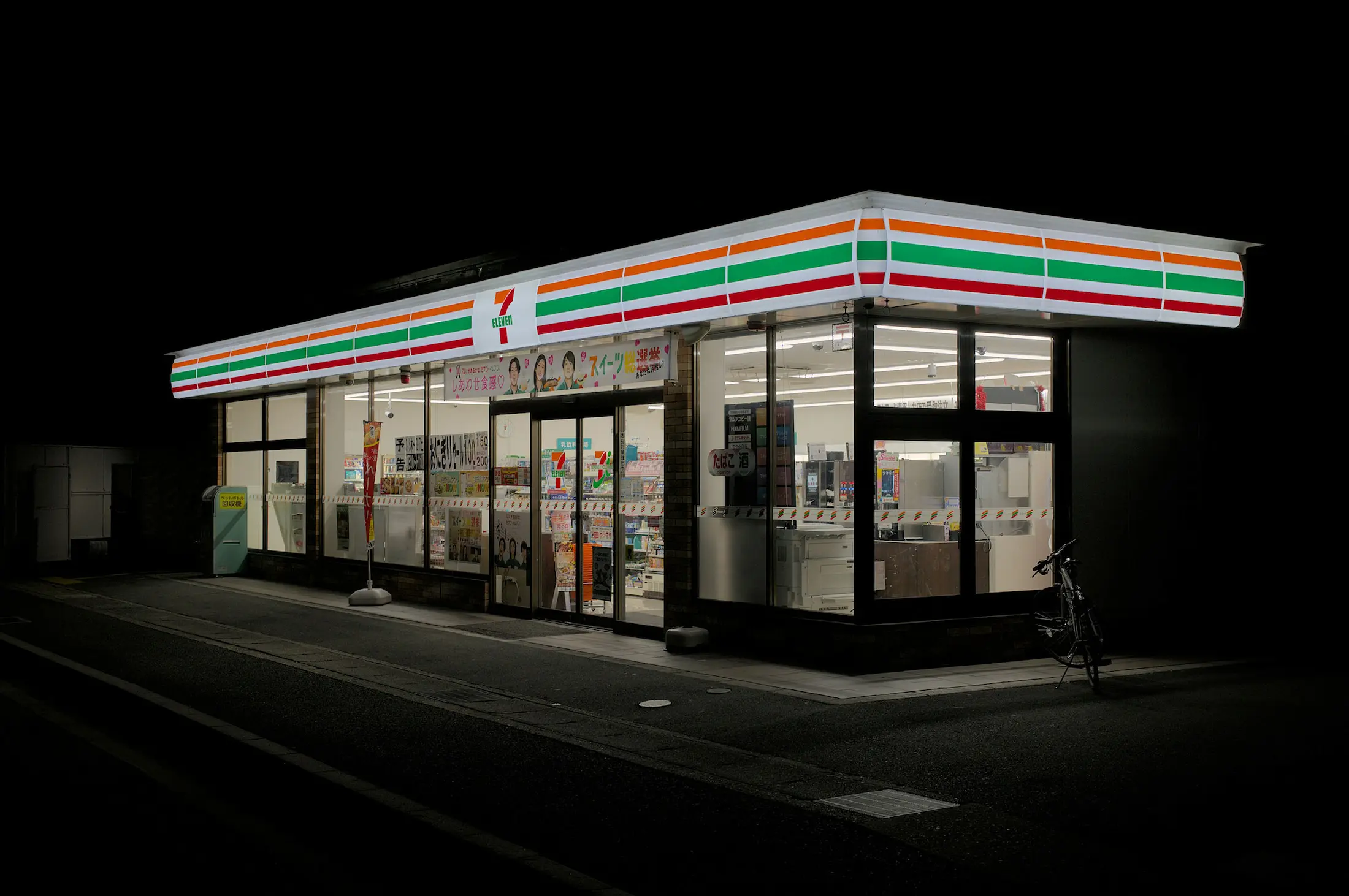 Fachada de una tienda Seven-Eleven en Japón iluminada por la noche, con el característico logo de franjas rojas y naranjas y el exterior brillante sobre la calle oscura